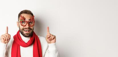 Christmas, New Year and celebration concept. Close-up of excited bearded guy in party glasses, pointing and looking up at store promo, white background photo