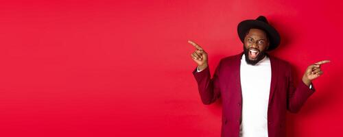 Winter holidays and shopping concept. Cheerful Black man smiling and showing two promo, pointing fingers sideways at copy spaces, red background photo