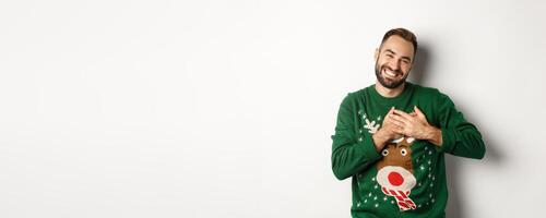 New year celebration and winter holidays concept. Happy young man looking grateful, holding hands on heart and thanking for christmas gift, standing in funny sweater photo