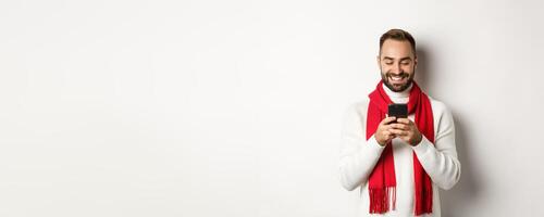 Happy man smiling while reading message on mobile phone, standing in winter sweater and red scarf against white background photo