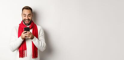 Man reading messaging on mobile phone and looking happy, standing in winter sweater and red scarf, white background photo