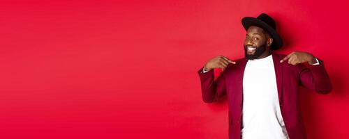 Christmas shopping and people concept. Handsome Black man pointing at himself, going on party, standing confident against red background photo
