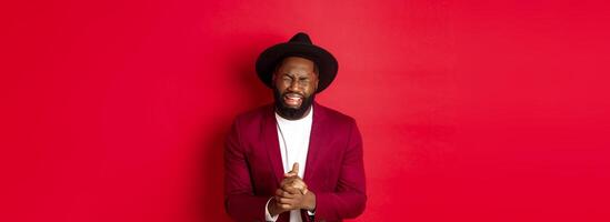 Miserable and heartbroken Black man crying, begging for help, holding hands in pray, standing against red background photo