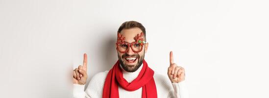 Christmas, New Year and celebration concept. Close-up of handsome bearded man in party glasses and red scarf, celebrate winter holidays and pointing fingers up at copy space photo