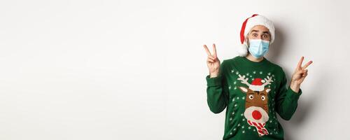 Concept of covid-19 and Christmas holidays. Happy man in medical mask and Santa hat showing peace signs, standing over white background photo