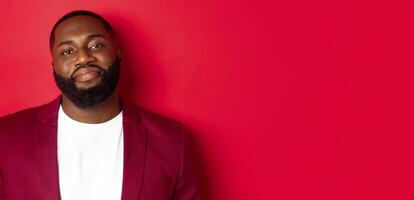 Close-up of handsome bearded plump guy smiling at camera shy, standing in stylish blazer on t-shirt, standing over red background photo