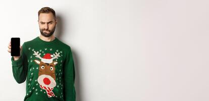 New Year, holidays and celebration. Skeptical guy looking doubtful at screen while showing it to you, demonstrating mobile app, standing over white background photo