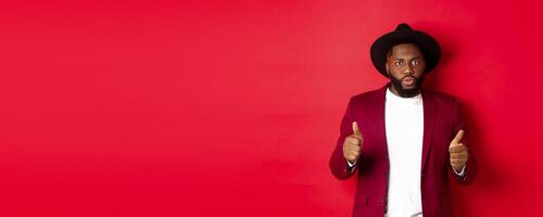 Impressed Black man showing thumbs up and staring amazed at camera, approve and praise something good, red background photo