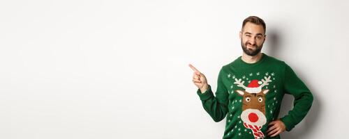 New year celebration and winter holidays concept. Skeptical guy in christmas sweater looking displeased, pointing finger at upper left corner product, dislike it, white background photo