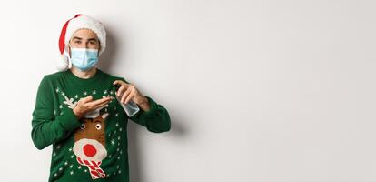 Concept of covid-19 and Christmas holidays. Caucasian man in face mask and sweater using antiseptic, clean hands with sanitizer, standing over white background photo