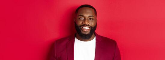 Close-up of handsome and stylish Black man wearing party blazer, smiling happy at camera, standing against red background photo