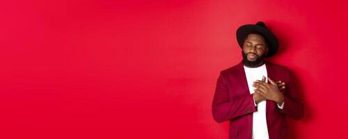 Fashion and party concept. Young bearded Black man in classy hat and jacket, holding hands on heart and smiling nostalgic, remember something, daydreaming against red background photo