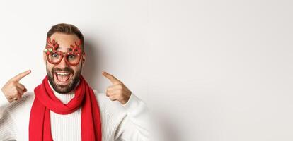 Christmas, New Year and celebration concept. Excited bearded man pointing at his party glasses, having fun and standing over white background photo