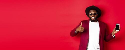 Handsome and stylish Black man showing phone screen and thumb up at camera, recommending online store app, red background photo