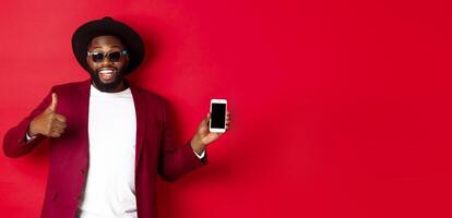 Handsome and stylish Black man showing phone screen and thumb up at camera, recommending online store app, red background photo