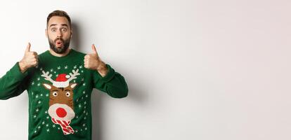 Christmas, holidays and celebration. Surprised young man in green sweater, showing thumbs up in approval, like something great, standing over white background photo