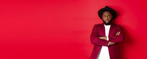 Reluctant and upset Black man waiting for apology, cross arms on chest and looking offended at camera, red background photo
