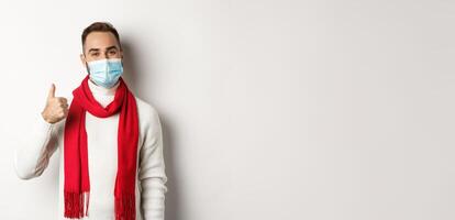 Covid-19, lockdown and quarantine concept. Satisfied smiling man in face mask showing thumb up, approve or like product, standing against white background photo