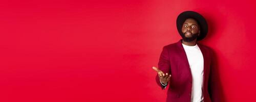 Shocked african american man staring at camera and pointing at something upsetting, standing in party jacket and hat, red background photo