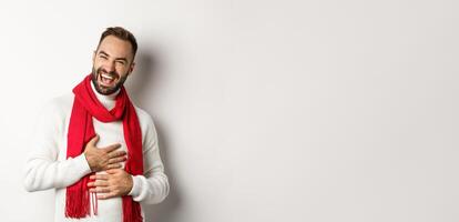 Happy man laughing and touching belly, chuckling from funny joke, standing in winter sweater and red scarf, white background photo