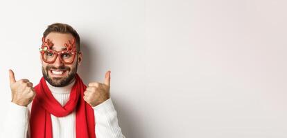 Christmas, New Year and celebration concept. Happy and satisfied man with beard, wearing party glasses, showing thumbs up in approval or like, standing over white background photo