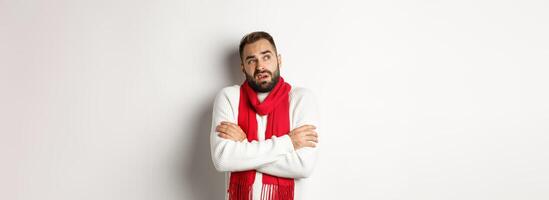 Confused man looking doubtful, standing with hands crossed and feeling cold on winter, wearing christmas sweater with red scarf, standing over white background photo