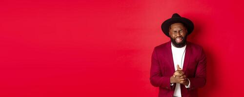 Miserable and heartbroken Black man crying, begging for help, holding hands in pray, standing against red background photo
