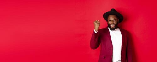 Fashion and party concept. Excited Black man rejoicing of winning, making fist pump gesture and say yes with satisfaction, standing against red background photo