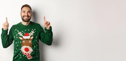 Winter holidays and christmas. Happy smiling man in funny sweater pointing fingers up, showing advertisement, standing over white background photo