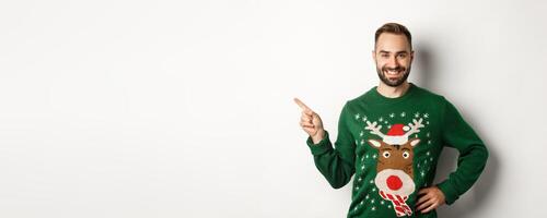 New year celebration and winter holidays concept. Confident and happy man with beard, wearing christmas sweater, pointing at upper left corner banner, white background photo