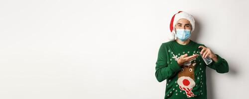 Concept of covid-19 and Christmas holidays. Caucasian man in face mask and sweater using antiseptic, clean hands with sanitizer, standing over white background photo