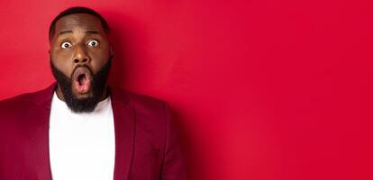 Close-up of shocked Black man gasping and dropping jaw impressed, staring at camera, standing over red background photo