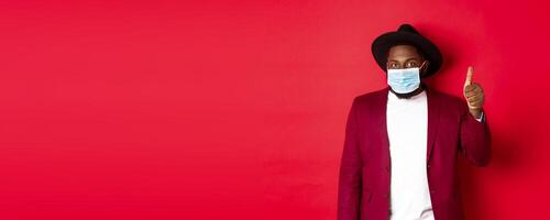 Covid-19 and fashion concept. Stylish african american man in hat and blazer, wearing face mask and showing thumb up, standing over red background photo