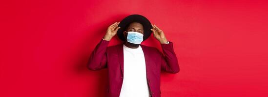 Covid-19, quarantine and holidays concept. Handsome and stylish african american man in face mask, put hat on head and looking sassy, dress-up for party, standing over red background photo