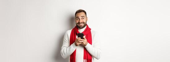 Man thinking while texting message on mobile phone, smiling and standing over white background photo