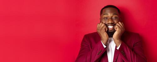 Close-up of silly african american man watching something adorable, touching cheeks and smiling with delight and happiness, standing over red background photo