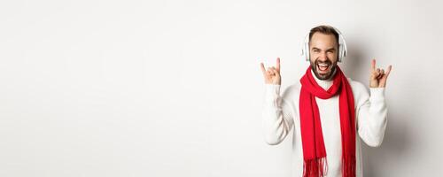 Joyful guy listening heavy metal in headphones, showing rock-on gesture and shouting for joy, standing over white background photo