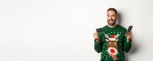 New Year, online shopping and christmas concept. Happy bearded man using mobile phone and credit card, winking at camera, order something in internet, white background photo