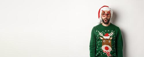 Christmas holidays, celebration concept. Surprised man in Santa hat and party glasses looking at something awesome, standing over white background photo