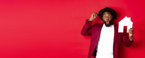 Real estate. Cheerful Black man showing paper house and smiling, recommending broker, standing over red background photo