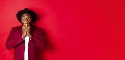 Fashion and party concept. Shocked Black man react on christmas advertisement, gasping and staring at camera with disbelief, standing over red background photo