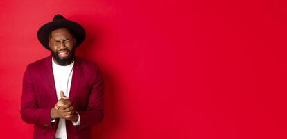Miserable and heartbroken Black man crying, begging for help, holding hands in pray, standing against red background photo