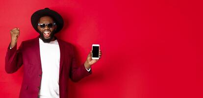 Cheerful winning Black man showing smartphone screen, rejoicing and demonstrating application, standing over red background photo
