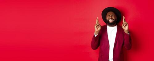 Hopeful african american man making wish, holding fingers crossed for good luck and smiling optimistic, standing against red party background photo