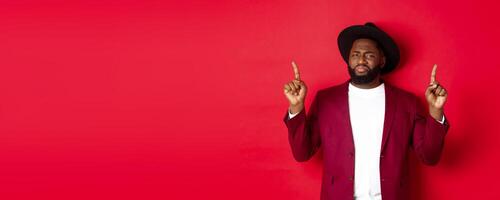 Winter holidays and shopping concept. Skeptical Black man grimasing and showing dislike, pointing fingers up at bad offer, standing over red background photo