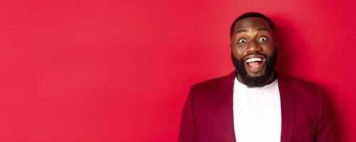 Close-up of african american man with beard staring amazed at camera, smiling and looking excited, standing in blazer against red background photo