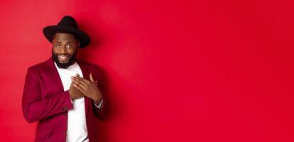 Fashion and party concept. Handsome african american macho man flirting, smiling and holding hands on heart, being grateful and touched, standing over red background photo