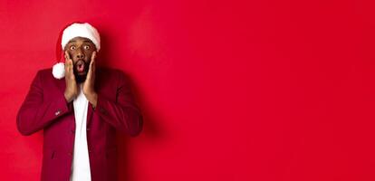 Christmas, party and holidays concept. Shocked Black man in santa hat reacting to new year offer, standing against red background photo