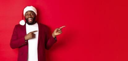 Christmas, party and holidays concept. Cheerful Black man smiling, pointing fingers right and showing advertisement, standing over red background photo