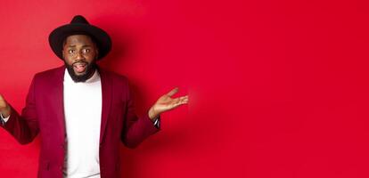Image of confused Black man asking question, spread hands sideways and staring camera clueless, standing against red background photo